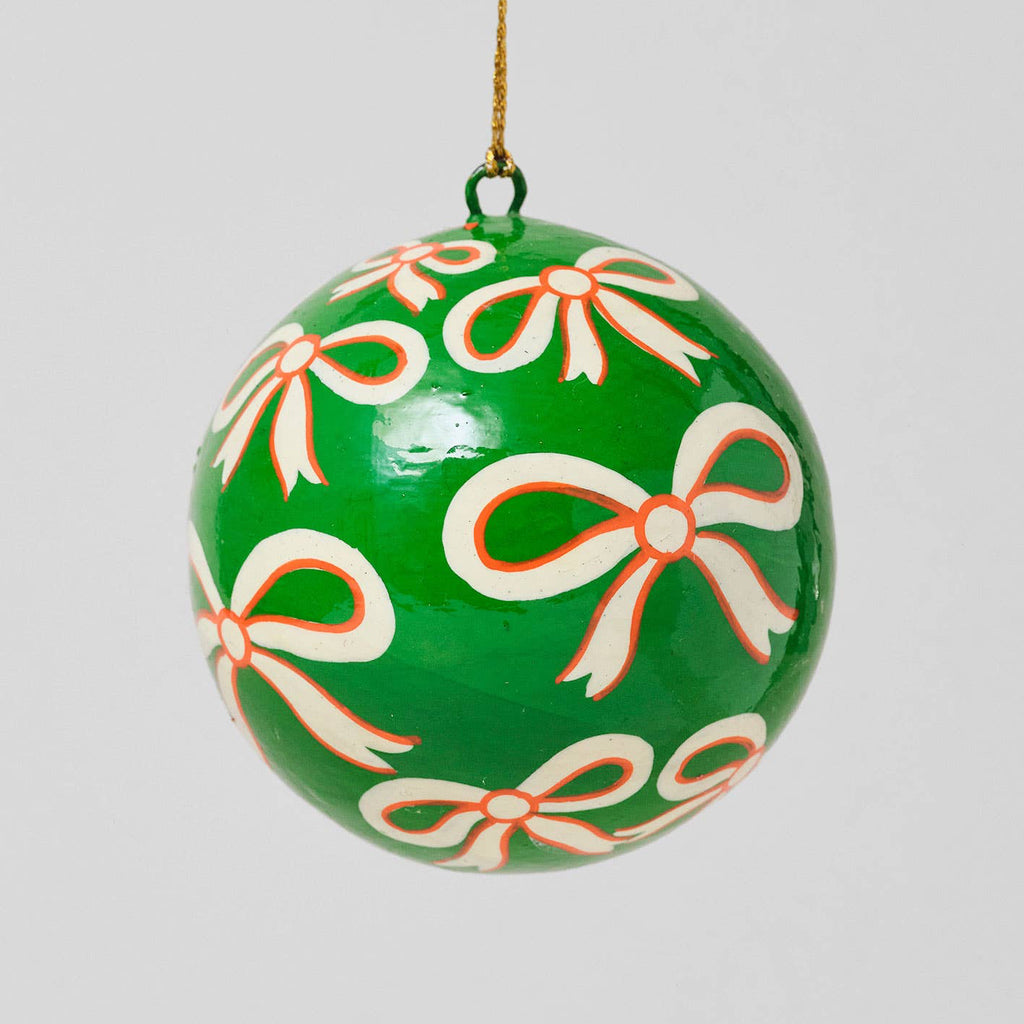 A green Christmas ornament shaped like a hanging bauble, decorated with a repeating pattern of white bows outlined in orange, suspended by a thin gold string against a light gray background.