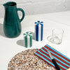 Green ceramic pitcher, striped candle, glass, and striped cutting board on a textured surface.