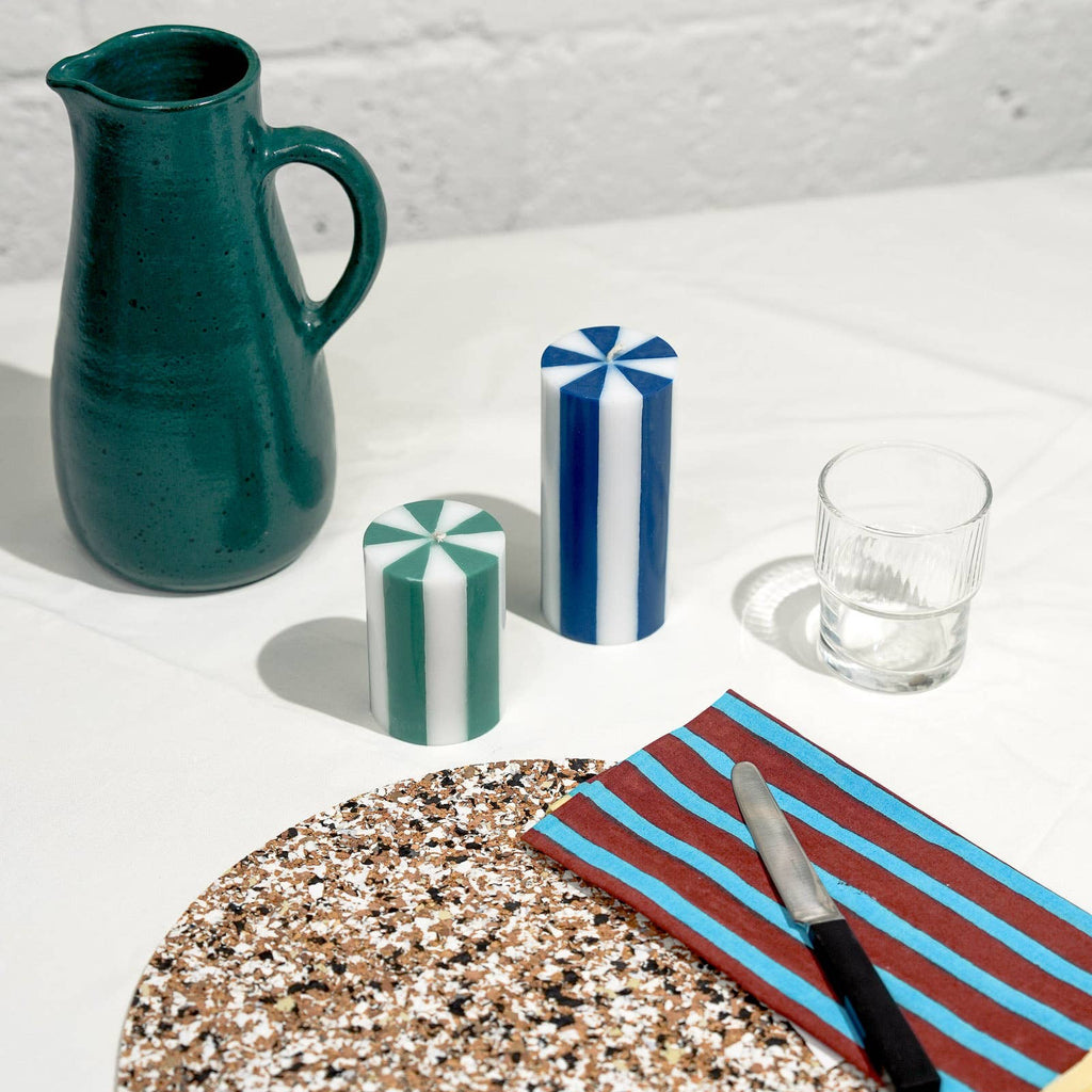 Green ceramic pitcher, striped candle, glass, and striped cutting board on a textured surface.