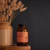 A bottle of Sweet Bee Organics Nutritious Balm sits on a wooden stool next to a vase with dried grasses, with a person's legs stretched out in a bathtub in the softly blurred background.