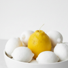 A single bright yellow lemon sits in the center of a white ceramic bowl filled with white lemons, photographed against a clean white background.