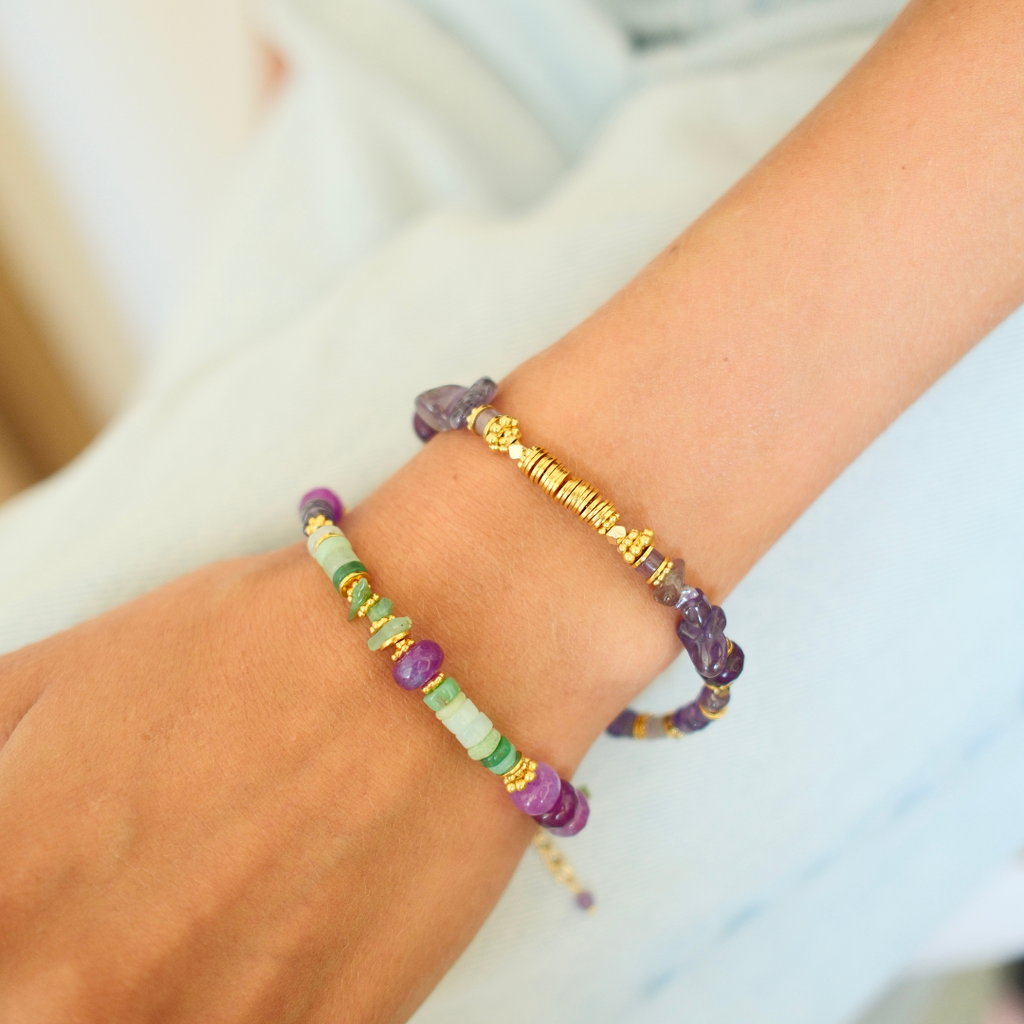 A close-up of a person's wrist wearing two beaded bracelets. One bracelet is made of purple translucent stone beads, and the other is made of gold-toned textured beads, both layered on a light skin tone against a soft white background.