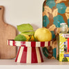 A red and cream striped pedestal fruit bowl filled with lemons and green leaves, styled on a shelf with a wooden cutting board, a patterned tray, and a bottle in the background.