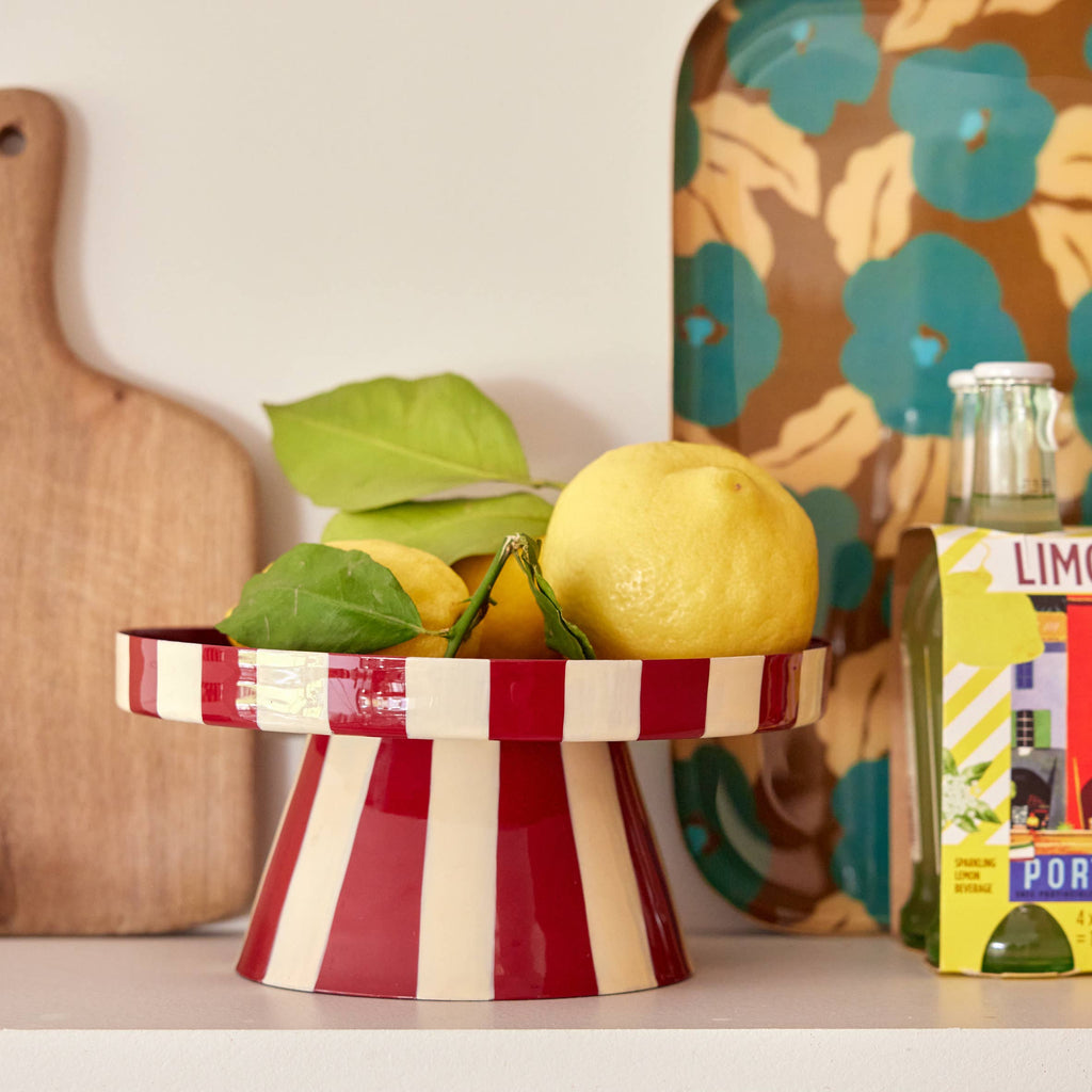 A red and cream striped pedestal fruit bowl filled with lemons and green leaves, styled on a shelf with a wooden cutting board, a patterned tray, and a bottle in the background.