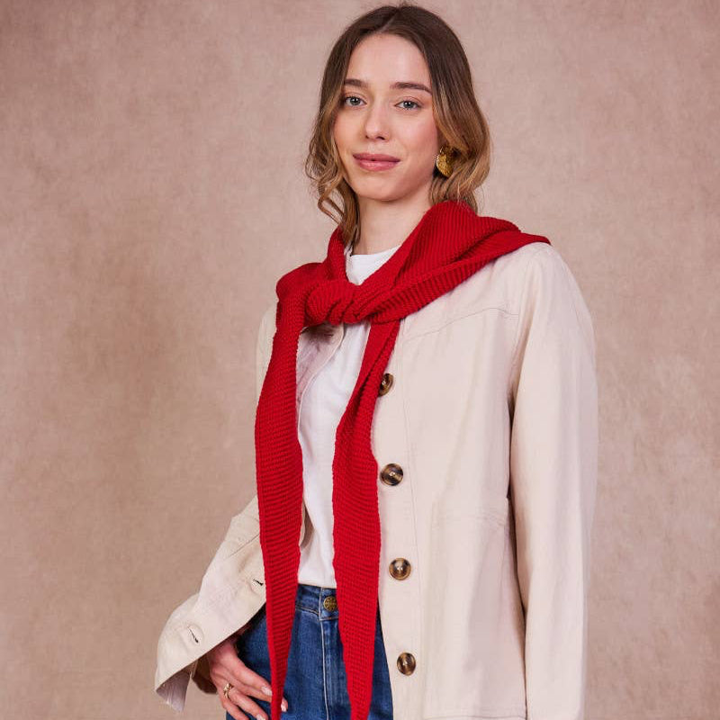 Woman wearing a red scarf, white shirt, beige jacket, and blue jeans against a beige background