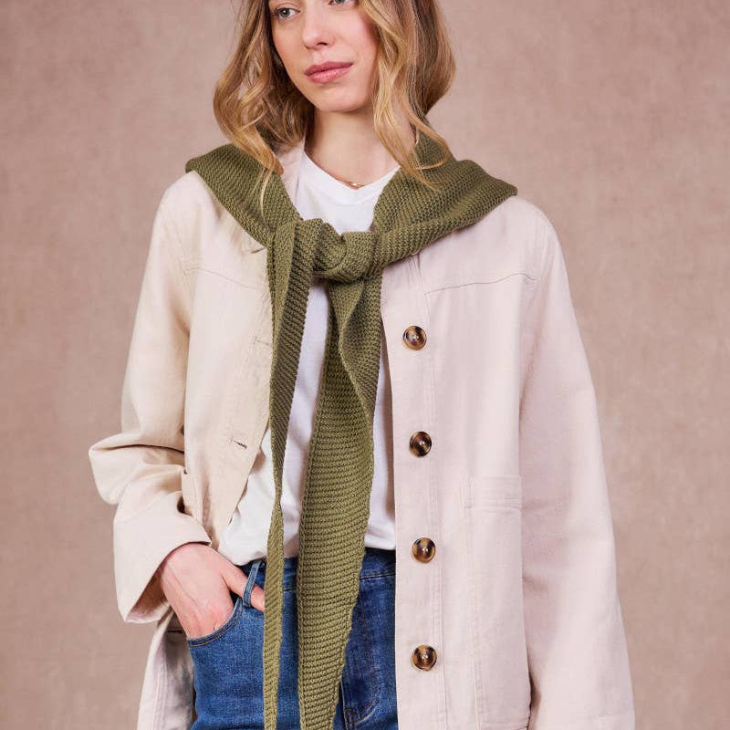 Woman wearing a beige coat, white shirt, blue jeans, and a green scarf against a beige background