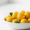 A white ceramic bowl filled with yellow citrus fruit candles, some with lit wicks and small green leaves, set against a clean white background.