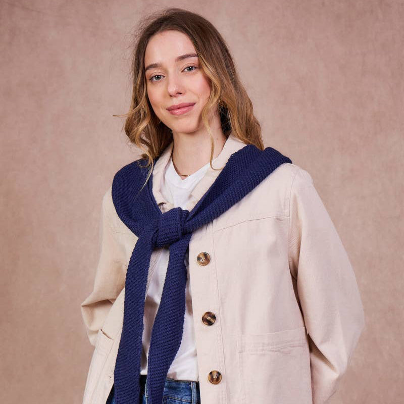Woman wearing a beige coat, blue scarf, and jeans against a pink background