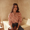 Woman in a patterned shirt and jeans holding a woven bag in a warm-toned room.