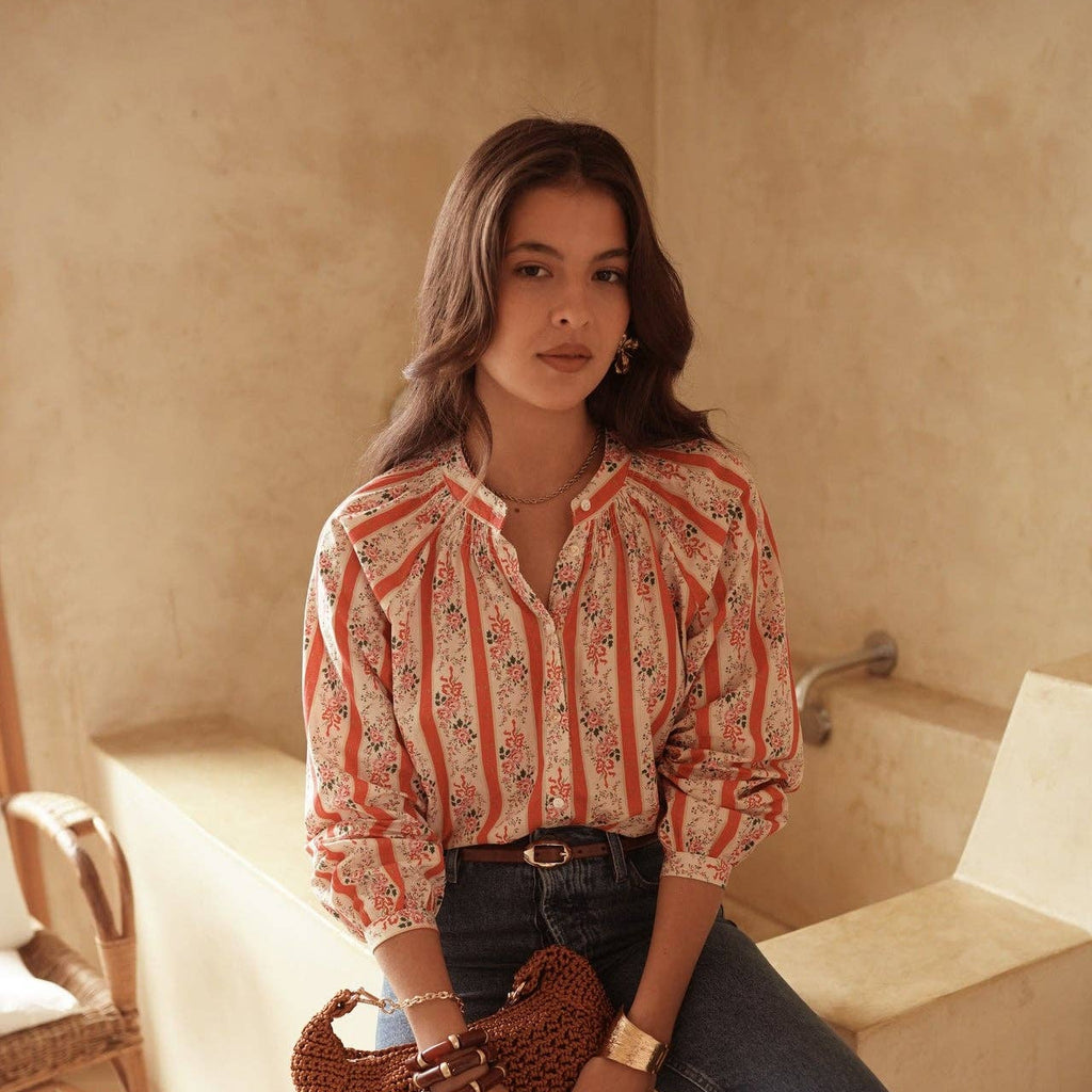 Woman in a patterned shirt and jeans holding a woven bag in a warm-toned room.