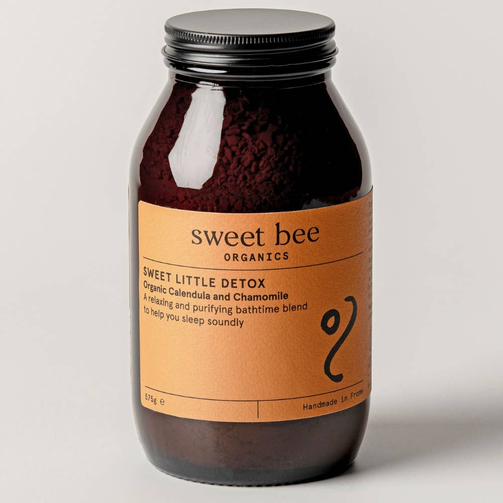 A dark amber glass jar of Sweet Little Detox bathtime blend by Sweet Bee Organics, featuring a black metal lid and a warm tan label, set against a neutral light gray background.