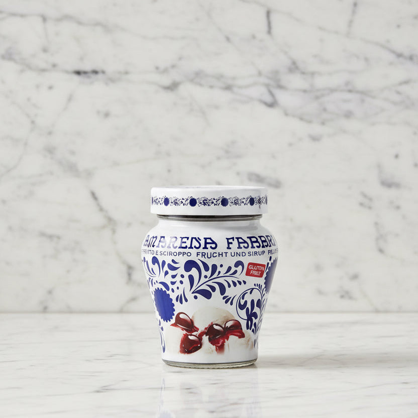 A glass jar of Amarena Fabbrica fruit and syrup, featuring a white label with blue floral patterns, German text, a red 'Gluten Free' badge, and an illustration of ice cream and dark red syrup.