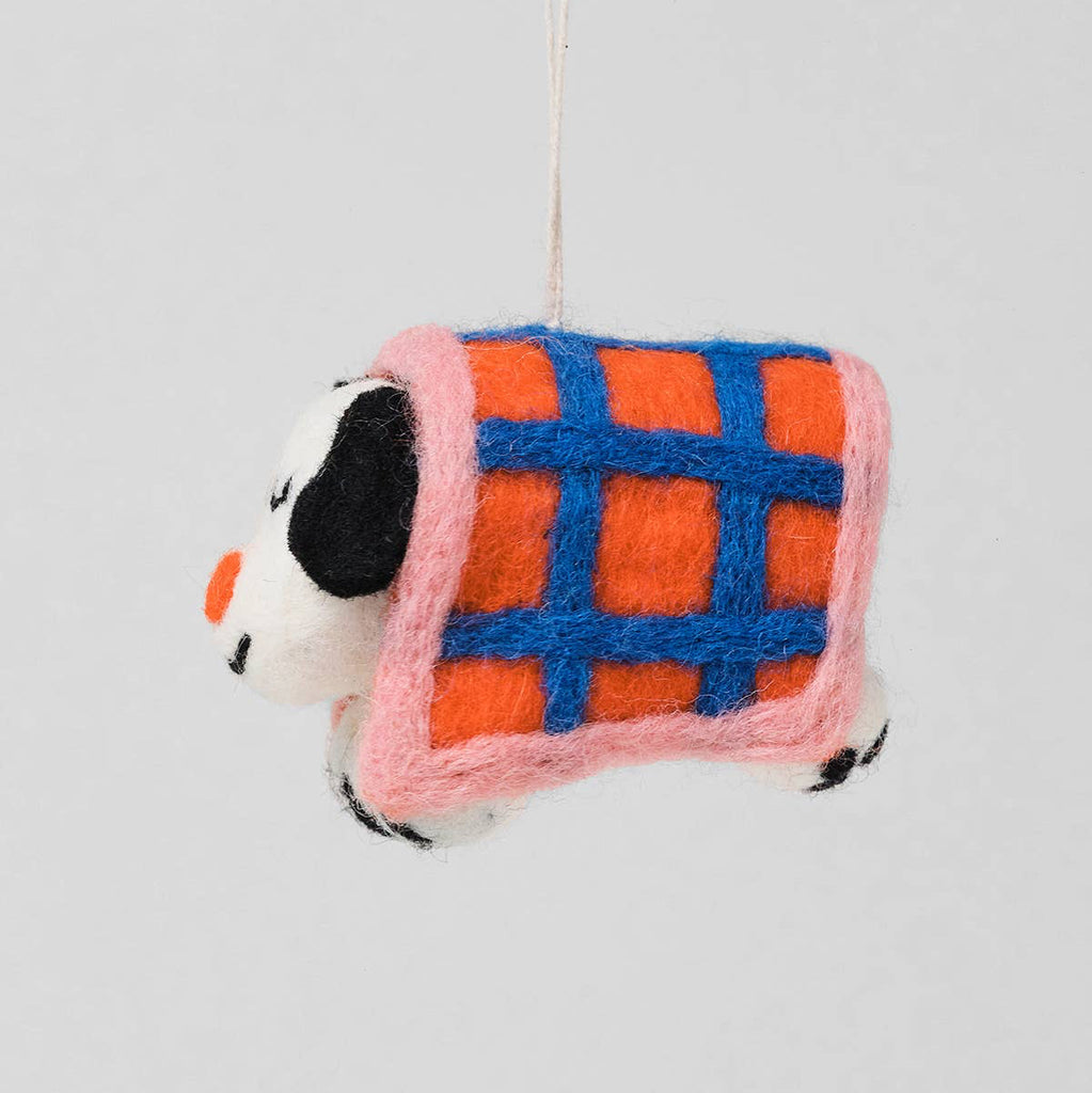A handcrafted felt dog ornament wrapped in a plaid blanket with orange, blue, and pink colors, hanging against a light gray background.