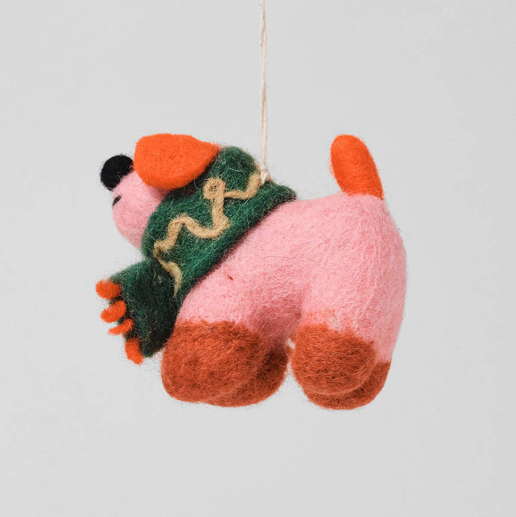 A pink felt dog hanging ornament wearing a green and beige patterned sweater, suspended by a thin string against a light gray background.