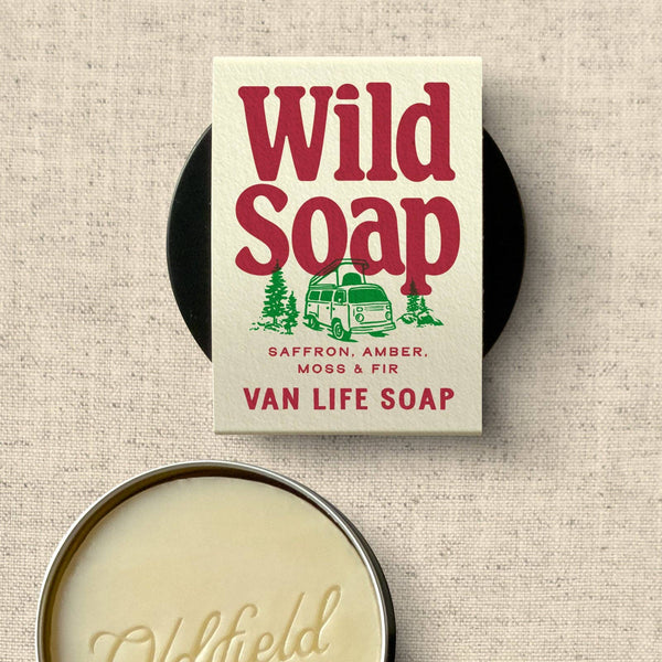A tin of Oldfield Van Life Soap shown open, with the cream-colored soap bar resting in front of the product box on a textured beige fabric background. The box features red and green retro-style lettering that reads 'Wild Soap' with a vintage camper illustration.