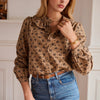A woman wearing a brown long-sleeve blouse with a black floral pattern, paired with blue jeans and gold jewelry, in an indoor setting with white walls.