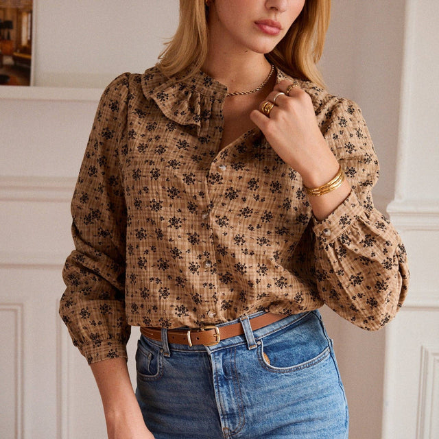 A woman wearing a brown long-sleeve blouse with a black floral pattern, paired with blue jeans and gold jewelry, in an indoor setting with white walls.