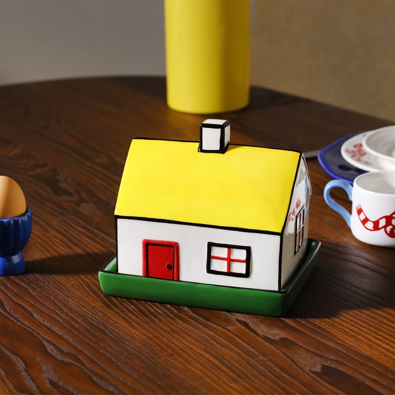 A ceramic butter dish designed to resemble a house, with a yellow roof, red door, and green windows, placed on a wooden surface.