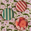 Three decorative round plates with colorful patterns are arranged on a green and pink floral fabric background. One plate has teal and white vertical stripes, one has red and white diagonal stripes, and one has large pink floral motifs on a coral-red background.