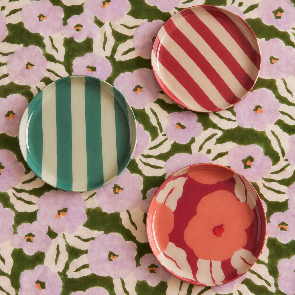 Three decorative round plates with colorful patterns are arranged on a green and pink floral fabric background. One plate has teal and white vertical stripes, one has red and white diagonal stripes, and one has large pink floral motifs on a coral-red background.