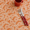 An orange tablecloth with a white floral pattern, styled with a matching orange mug and a set of red-handled flatware on a light surface.