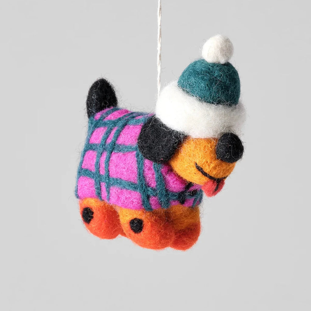 A felted wool dachshund hanging ornament wearing a pink and teal plaid sweater, a teal and white beanie hat, and orange paws, suspended by a white string against a solid gray background.