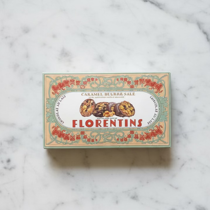 A vintage-style box of Florentins caramel-filled brioche cookies placed on a distressed, weathered concrete floor. The box features an ornate cream-colored label with red floral borders and the brand name 'FLORENTINS' printed in red.
