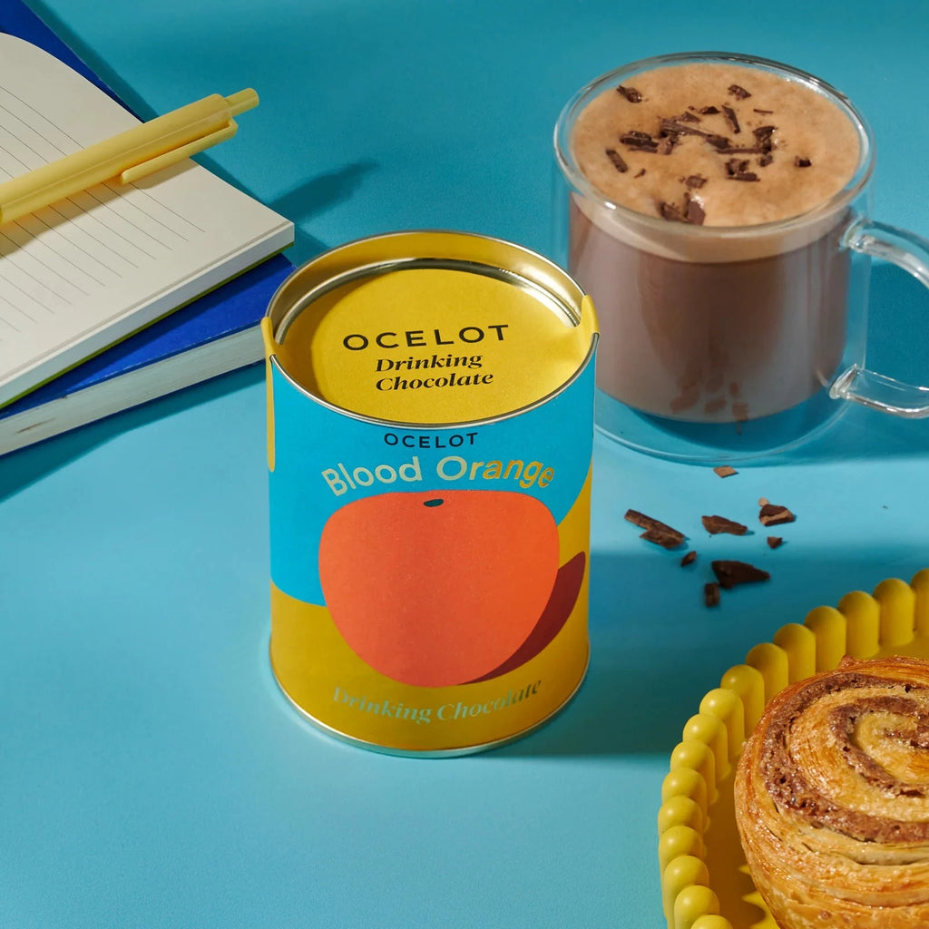 Ocelot Blood Orange drinking chocolate can with a cup of hot chocolate and a pastry on a blue background