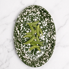 An oval dark green enamel plate with white speckled pattern, containing green pea pods, set against a solid pink background.