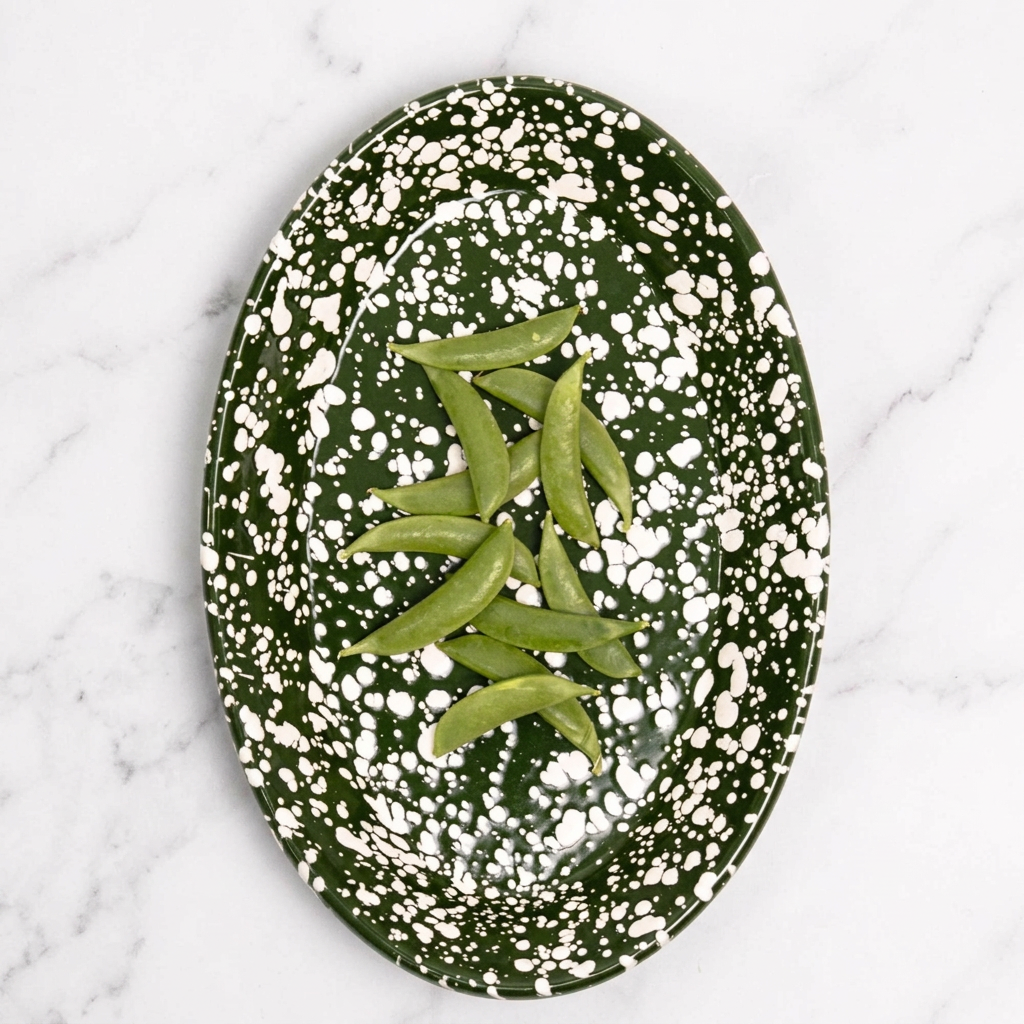 An oval dark green enamel plate with white speckled pattern, containing green pea pods, set against a solid pink background.