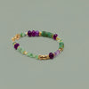 A beaded stretch bracelet featuring green and purple gemstone beads, gold spacer beads, and a gold clasp, displayed on a clean white background.