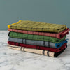 A stack of six folded plaid blankets in various colors, including olive green, dusty rose, forest green, slate blue, burgundy, and cream, set against a plain light gray background.