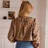 A woman viewed from behind wearing a brown and black floral patterned blouse with puff sleeves and blue jeans, standing in a bright indoor room with white walls and shelves.