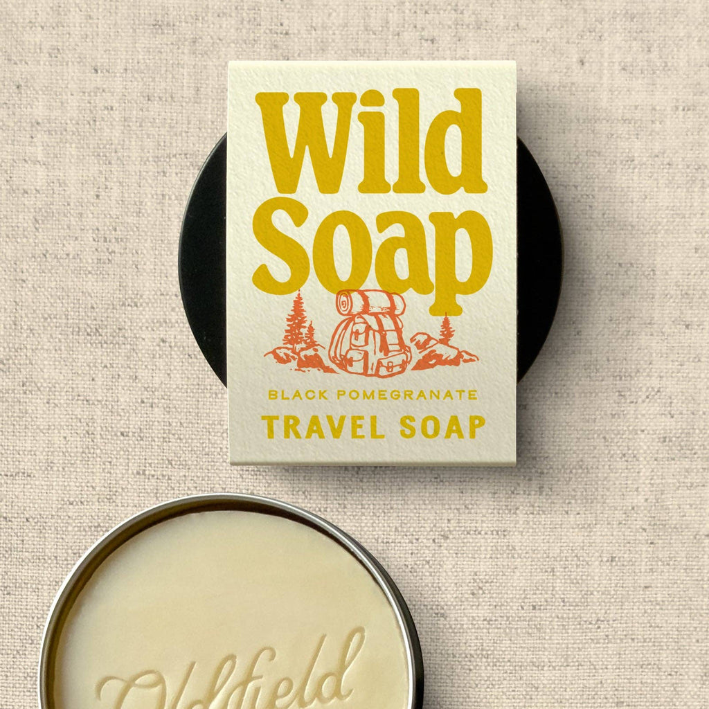 A travel soap product shown on a textured beige fabric background, with the front label reading 'Wild Soap' and 'Black Pomegranate Travel Soap,' plus a round metal tin of cream-colored soap below it.