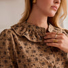 Close-up of a woman wearing a brown crinkled blouse with a black floral print, featuring a ruffled collar and button front.