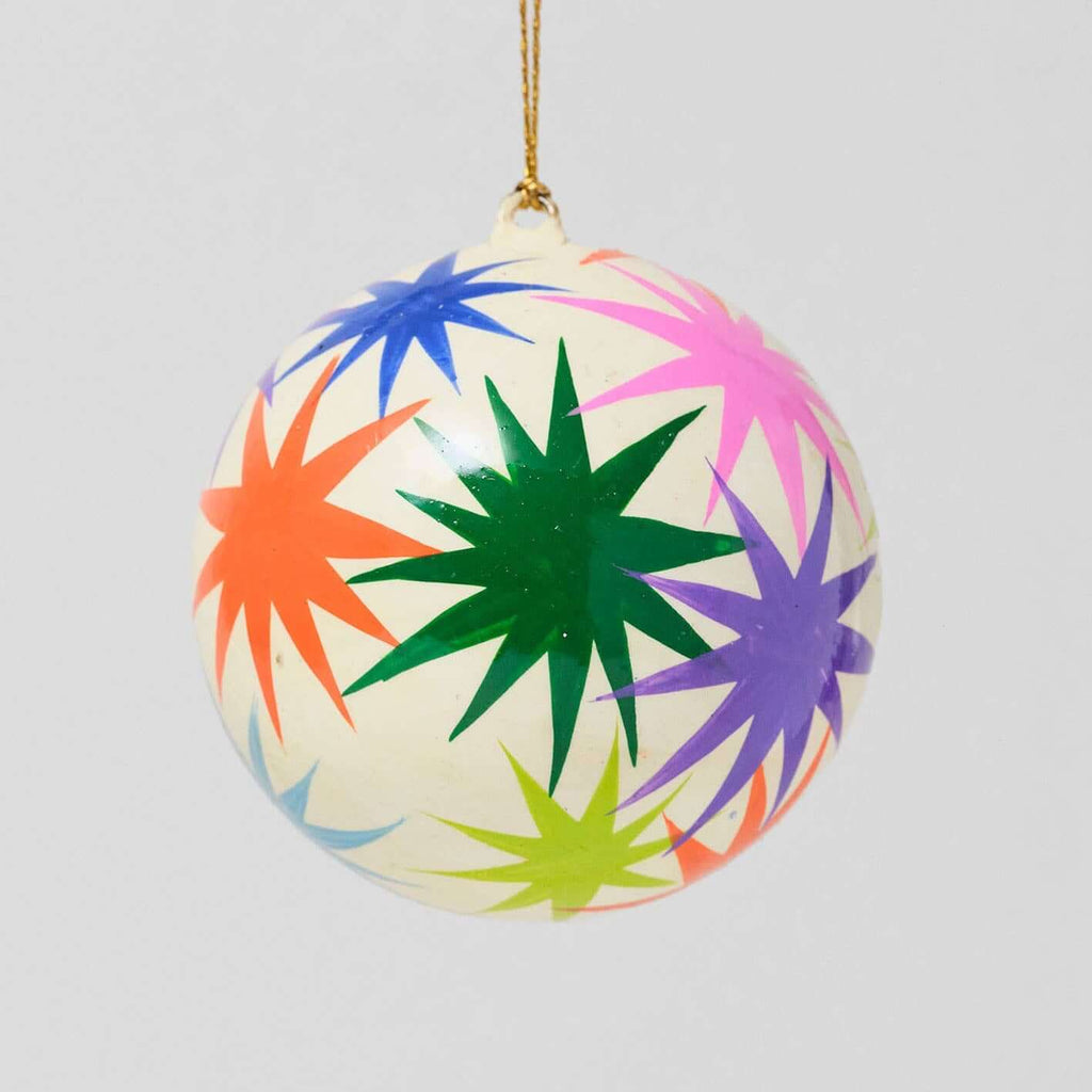 A spherical Christmas ornament with a glossy cream background and a colorful hand-painted starburst pattern in green, purple, pink, orange, blue, and yellow, hanging by a gold string against a light gray background.