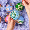 A close-up of hands holding a light blue hand-painted Christmas ornament with green floral patterns, surrounded by other colorful hand-painted ornaments on patterned purple and white fabric.