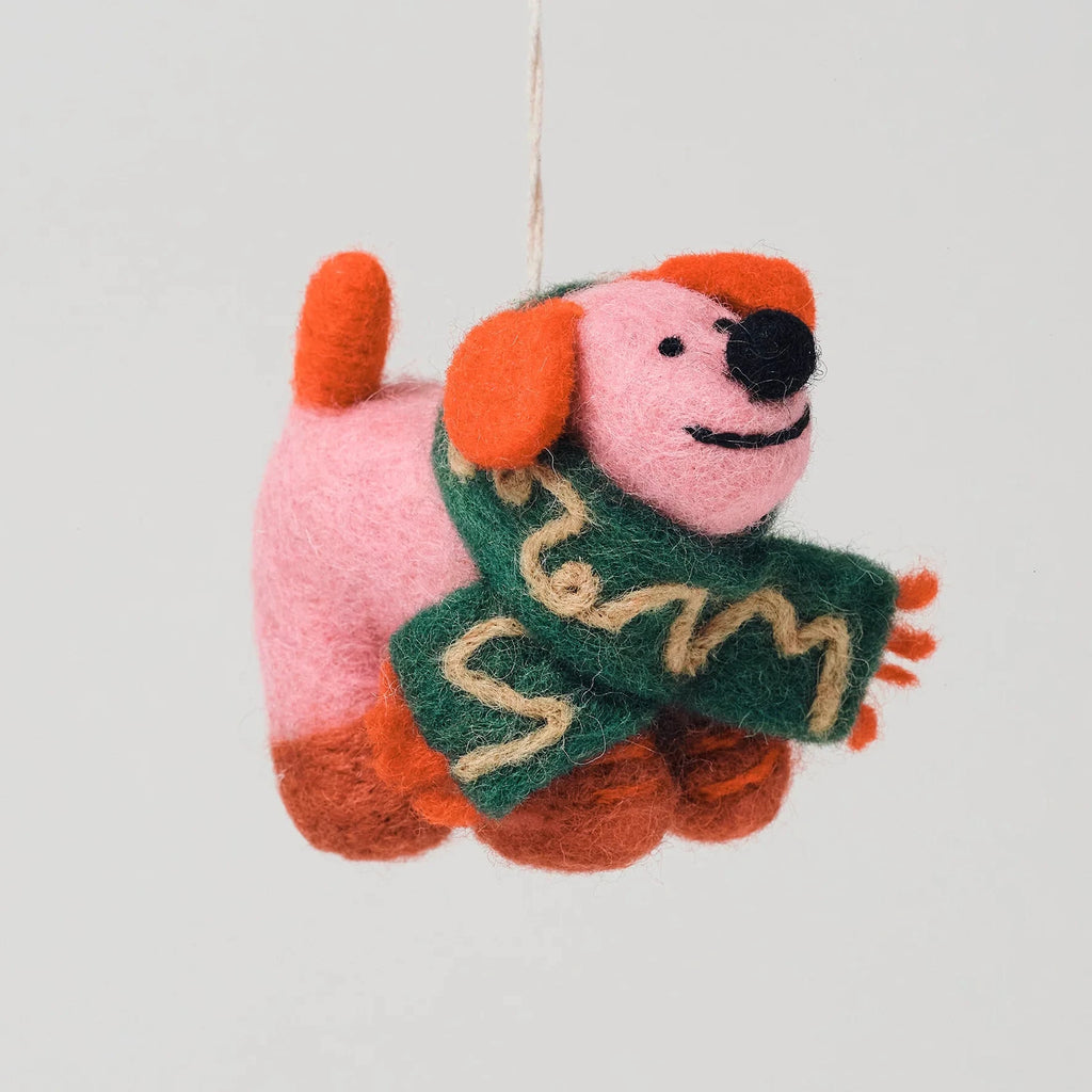 A felted wool dog ornament hanging from a thin string against a solid light gray background. The dog features a pink body, orange ears and paws, a green scarf with beige patterns, and a black nose.