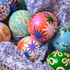 A close-up of colorful hand-painted Christmas ornaments nestled in patterned white and purple tissue paper inside a white basket. The ornaments feature vibrant colors like orange, green, blue, pink, and cream with hand-drawn starburst and bow patterns.