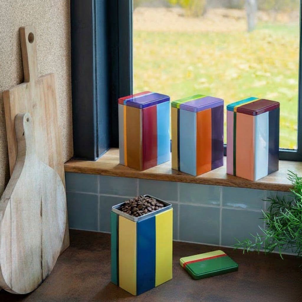 Colorful cookie tins on a windowsill with cookies on a cooling rack below.