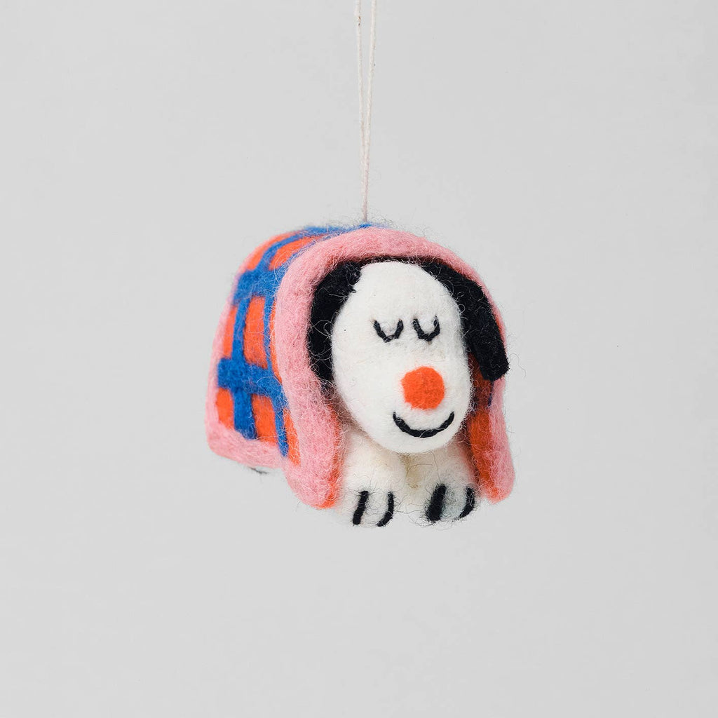 A felt dog ornament wrapped in a pink blanket with blue and orange plaid stripes, hanging against a light gray background.