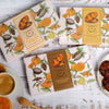 Candle boxes with orange and leaf design on a rustic wooden surface with cookies and coffee.