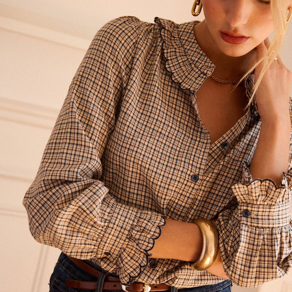 A woman wearing a beige and brown plaid button-down blouse with ruffled trim, tucked into dark blue jeans with a brown belt.