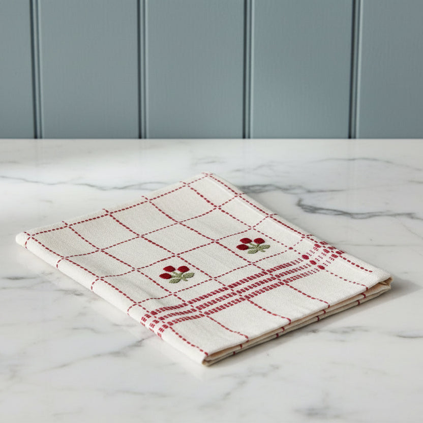 An off-white linen tea towel with a red stitched grid pattern, a red and green floral motif, and a folded corner showing the reverse side, set against a plain white background.