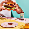 Person holding a burger with a bottle of hot sauce and glass of beer on a colorful background