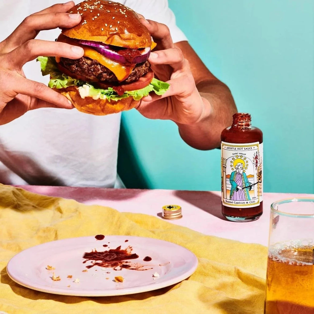 Person holding a burger with a bottle of hot sauce and glass of beer on a colorful background