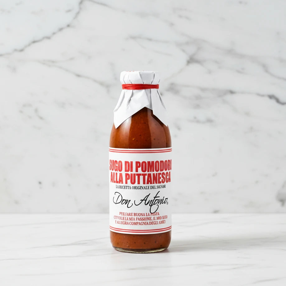 A glass bottle of Don Antonio 'Sugo di Pomodoro alla Puttanesca' sauce, featuring a white label with red and black text, topped with a white cloth and red ribbon, on a white background.