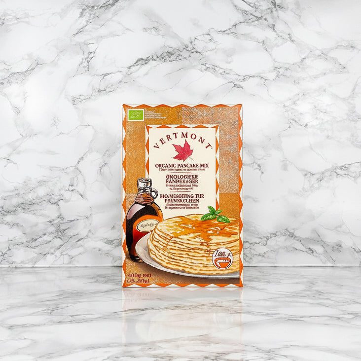 A package of Vertmont Organic Pancake Mix stands upright on a white marble surface against a matching white marble background. The packaging is orange and white with a photo of pancakes topped with syrup and a bottle of maple syrup on the front.